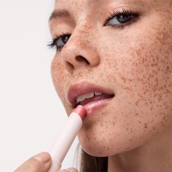 Kobieta z piegami nakłada błyszczyk Catrice Diamond Glaze Stick Glow For It, szkliste, koralowe usta.