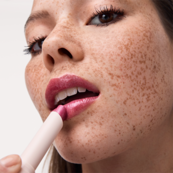 Zbliżenie: kobieta aplikuje błyszczyk Catrice Diamond Glaze Gloss Stick w jagodowym kolorze, z mokrym wykończeniem.