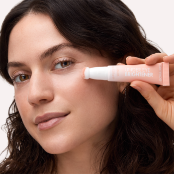 Kobieta aplikuje jasnoróżowy korektor rozświetlający Under Eye Brightener z gąbkowym aplikatorem pod oko.