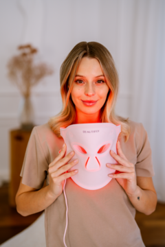 Kobieta trzyma różową maskę LED Beautifly Aura Glam PRO z aktywnym czerwonym światłem do fototerapii skóry.
