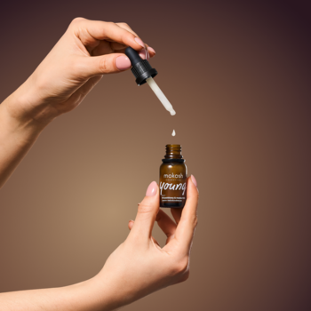 Kobieca dłoń trzyma pipetę z serum nad brązową buteleczką, skąd spada kropla płynu do pielęgnacji twarzy.