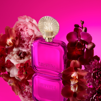 Różowa woda perfumowana Shakira Fucsia, kwadratowy flakon z promienistą nakrętką, wśród kwiatów.