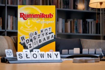 Rummikub Eco Słowny, gra słowna: widok na tekturowe kostki z literami "SŁOWNY" na szarej podstawce, pudełko w tle.