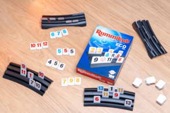 Rummikub Eco na drewnianym blacie: tekturowe kostki z liczbami, 3 czarne podstawki i niebieskie pudełko z grą.