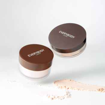 Dwa sypkie pudry rozjaśniające pod oczy Brightening Silk, z brązowymi zakrętkami, unoszą się nad smugami pudru.