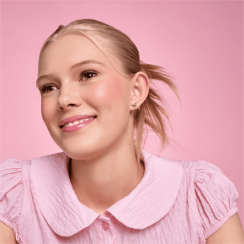Portret uśmiechniętej blondynki w różowej koszuli, z naturalnym różowym makijażem, na jasnoróżowym tle.