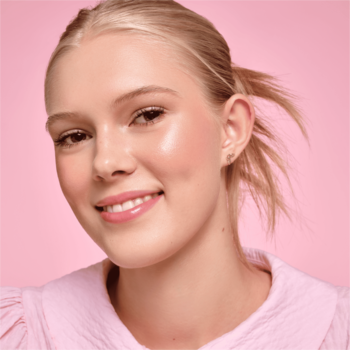 Uśmiechnięta blondynka z makijażem podkreślającym naturalny blask skóry i różowe usta, na różowym tle.