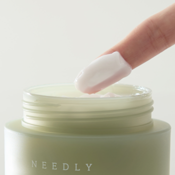 Palec z białą, budyniową konsystencją NEEDLY Cicachid Relief Cream, nabierający krem z jasnozielonego słoiczka.