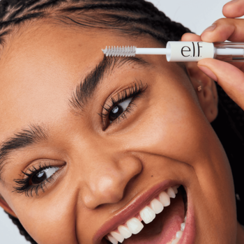 Uśmiechnięta kobieta z warkoczami aplikuje transparentny żel e.l.f. Clear Brow & Lash Mascara na brew. Zbliżenie.