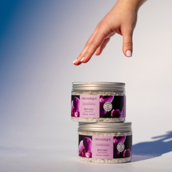 Dwa słoiki relaksującej soli do kąpieli Organique Black Orchid na stosie, z dłonią delikatnie dotykającą górnego.