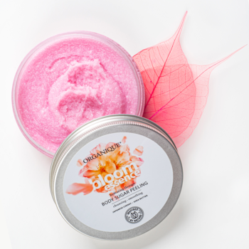 Organique Bloom Essence peeling cukrowy. Otwarty słoik z różowym scrubem, obok zamknięty, różowe liście na białym tle.