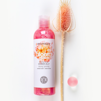 Organique Bloom Essence, różowy żel pod prysznic w butelce, obok suszona czerwona roślina ostu i mały różowy koralik.