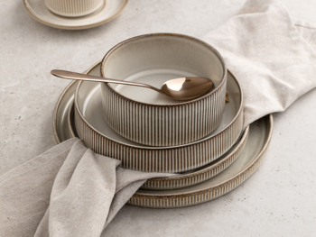 Jasnobeżowy zestaw naczyń ceramicznych z prążkowaniem: miska i talerze, ze złotą łyżką na lnianej serwetce.