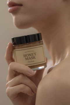 Nawilżająca maska I'm From Honey Mask w bursztynowym słoiku, trzymana przy ustach. Widok z przodu, kremowy składnik.