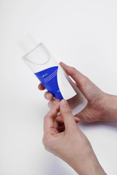Dłonie odklejają białą etykietę od butelki toniku Isntree Hyaluronic Acid Plus, pokazując niebieskie logo produktu.