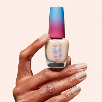 Dłoń z beżowym manicure trzyma szybkoschnący lakier OPI RapiDry Quick Sand, butelka z barwną zakrętką.