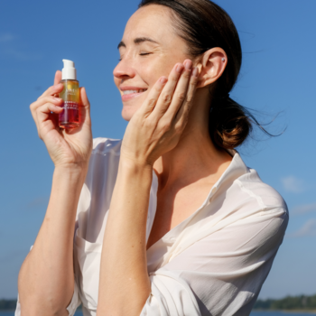 Kobieta aplikuje dwufazowy rozświetlający koktajl Lumene Nordic-C Valo z buteleczki. Uśmiechnięta na tle wody.