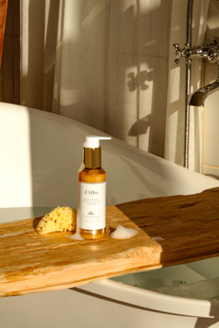 Biała butelka d'Alba White Truffle Oil Cream Cleanser z złotą pompką, na drewnianej półce w wannie z gąbką.