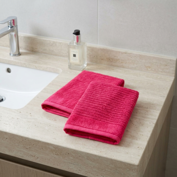 Dwa różowe bawełniane ręczniki łazienkowe, złożone na beżowym blacie obok białej umywalki.