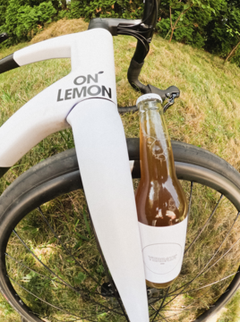 Napój ON° LEMON w butelce, przymocowany do białego widelca roweru, widok z bliska na tle zielonej trawy.