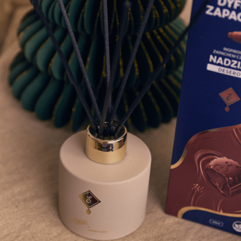 Biały dyfuzor zapachowy E. Wedel Nadziana deserowa z czarnymi patyczkami, obok opakowania czekolady.