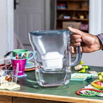 BRITA Style Essential, przezroczysty dzbanek filtrujący z szarym uchwytem i białym wkładem, trzymany ręką w kuchni.