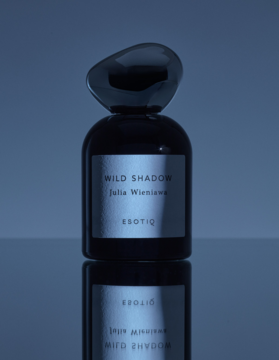 Ciemne perfumy Wild Shadow ESOTIQ z nieregularnym korkiem, srebrna etykieta Julia Wieniawa na odbijającej powierzchni.