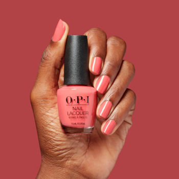 Dłoń trzyma butelkę koralowego lakieru OPI. Pomalowane paznokcie błyszczą, na czerwono-różowym tle.