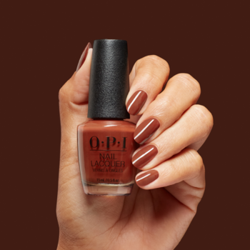 Ręka trzyma brązowy lakier do paznokci OPI Nail Lacquer z czarną nakrętką, paznokcie pomalowane tym kolorem.