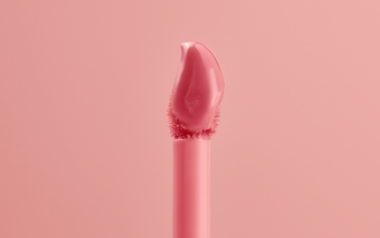 Zbliżenie na aplikator balsamu do ust z brzoskwiniowo-różową, błyszczącą formułą na jednolicie jasnoróżowym tle.