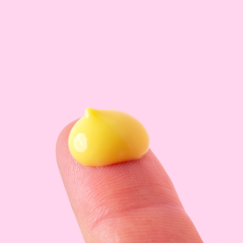 Serum A-Game 5 z retinalem 0.05%, mała żółta kropla emulsji na opuszku palca, różowe tło.