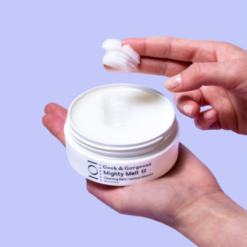 Biały słoik Geek & Gorgeous Mighty Melt Cleansing Balm, dłoń nabiera kremowy balsam do demakijażu.