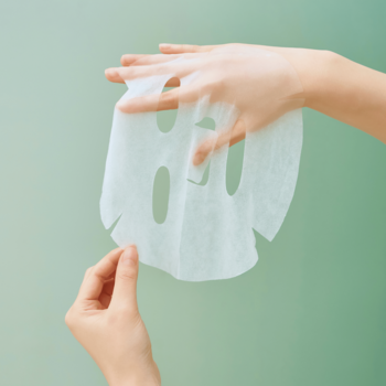 Dłonie rozkładające białą maskę w płachcie SKIN1004 Centella Tea-Trica Relaxing na zielonym tle.