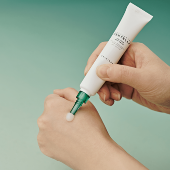 Ręka aplikuje Skin1004 Tea-Trica Spot Cream z białej tubki z zieloną nakrętką na dłoń, miętowe tło.