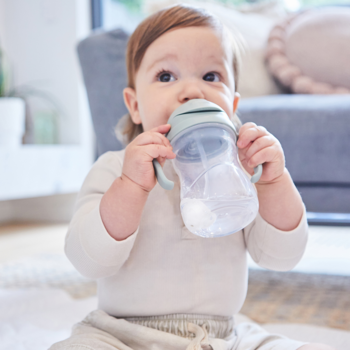 Dziecko pije wodę z zielonego bidonu b.box z uchwytami, siedząc na jasnym dywanie. Bidon ma obciążoną słomkę.