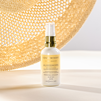 Sensum Mare Algodrops Satin SPF 50+ emulsja przeciw fotostarzeniu w białej butelce na tle kapelusza.