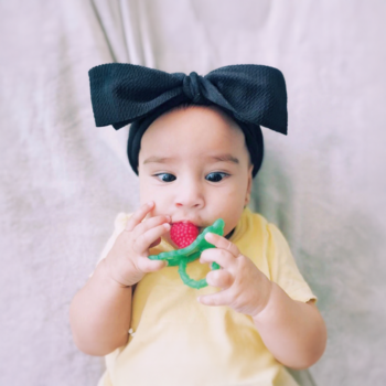 Dziecko w żółtej bluzce i czarnej kokardzie na głowie, leżące, przygryza silikonowy gryzak RaZbaby malinka.