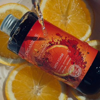 Organique Spicy Therapy Bath Foam, butelka z pomarańczową etykietą, lany płyn, otoczona świeżymi plastrami pomarańczy.