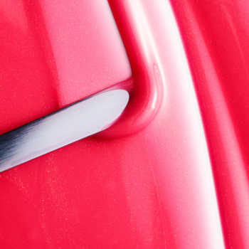 Zbliżenie na błyszczący, różowy balsam do ust Fluff Candy Lip Balm Crumbl Cookie, aplikowany srebrną szpatułką.