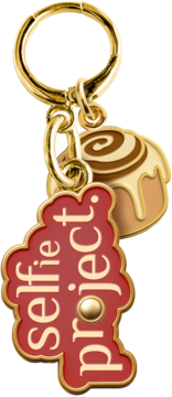 Złoty brelok z dwoma zawieszkami: cynamonką z polewą i czerwoną plakietką 'selfie project', z bliska.