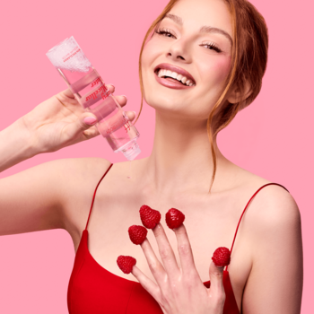 Kobieta trzyma płyn micelarny Fluff Berry do demakijażu, z malinami na palcach. Różowe tło.