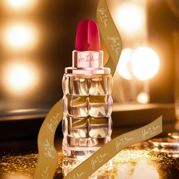 Perfumy Cacharel Yes I Am w złoto-czerwonym, szminkowym flakonie ze wstążką, na błyszczącym, ciepłym tle.