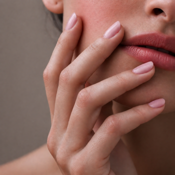 Ręka z paznokciami pomalowanymi lakierem COOLNAILS Blushing Hour w jasnym różu, dotykająca zarumienionego policzka.