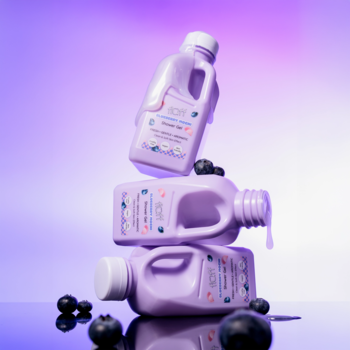 Trzy fioletowe butelki żelu pod prysznic Fluff Blueberry Mochi w stosie, ze świeżymi borówkami na fioletowym tle.