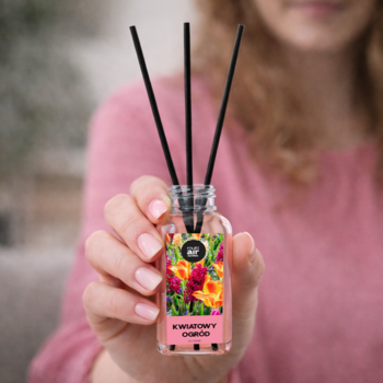 Dyfuzor zapachowy z patyczkami w szklanej butelce, czarne patyczki, etykieta "Kwiatowy Ogród", trzymany w dłoniach.