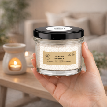 Ręka trzyma świecę zapachową Cotton Vanilla w szklanym słoiku z czarną pokrywką. Granulowany wosk, ciepłe tło.