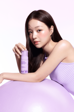 Kobieta trzyma fioletową butelkę SOME BY MI Retinol z pompką, oparta o fioletową piłkę, na białym tle.