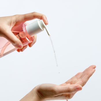 Różowy płyn kosmetyczny dozowany z przezroczystej butelki z pompką na otwartą dłoń.