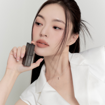 Zbliżenie na kobietę z matową pomadką Muzigae Mansion Nuddy, trzymającą sztyft przy ustach z efektem soft blur.