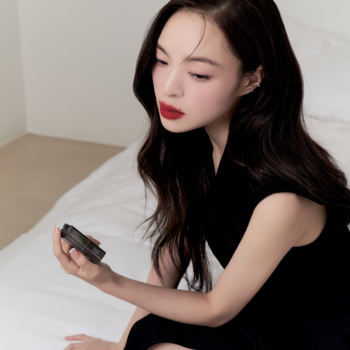 Kobieta z długimi włosami i czerwoną pomadką ogląda w dłoni matową pomadkę Muzigae Mansion Revenge, siedząc na białym łóżku.